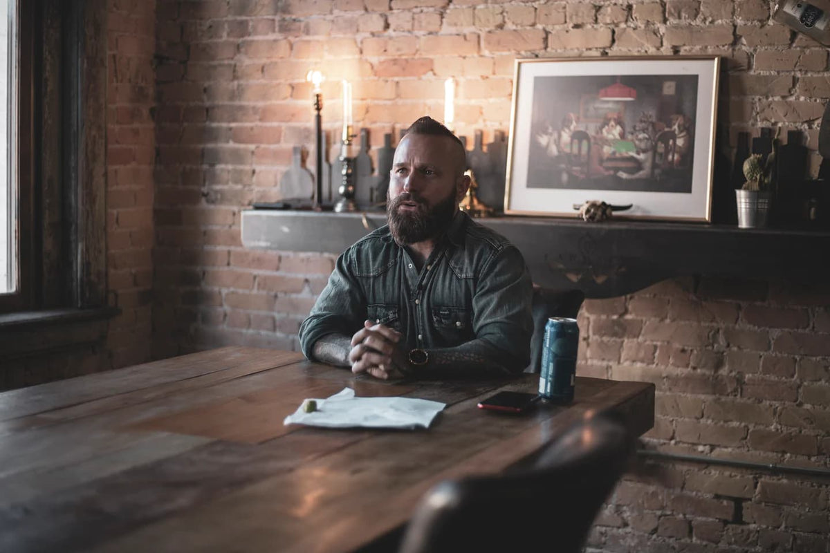 A bearded man in a denim shirt sits at a wooden table in a brick room